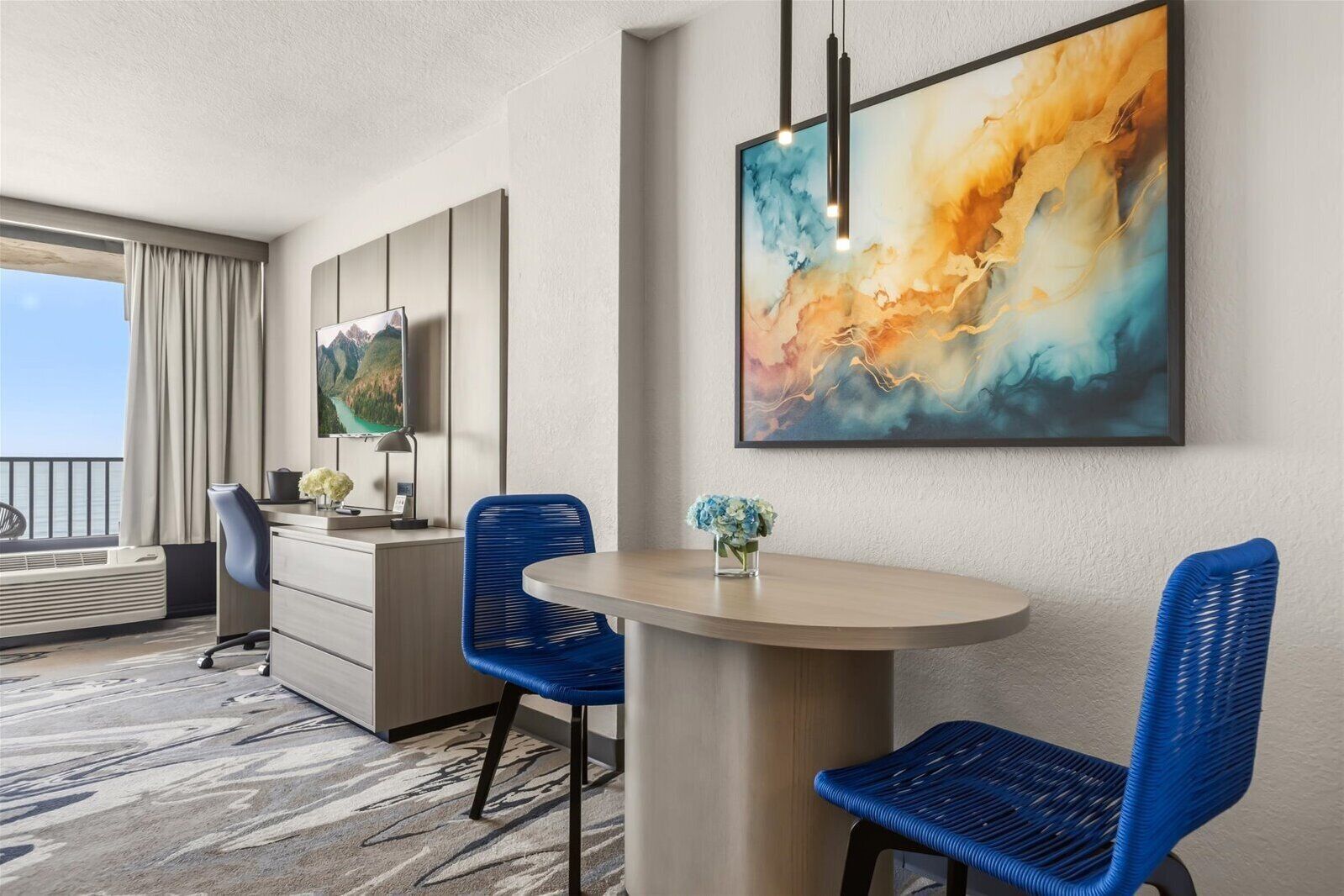 Habitación de hotel con zona de comedor, escritorio, silla, un florero con flores y un cuadro abstracto grande en la pared.