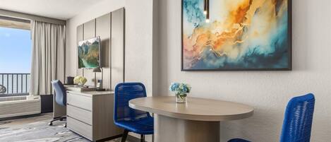 Habitación de hotel con zona de comedor, escritorio, silla, un florero con flores y un cuadro abstracto grande en la pared.