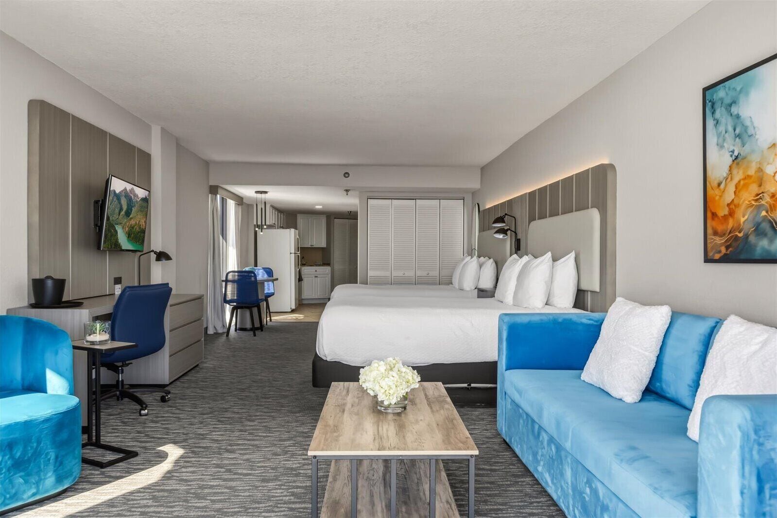 A hotel room with a bed, a blue sofa, a desk, a chair, a TV, and a kitchen area.