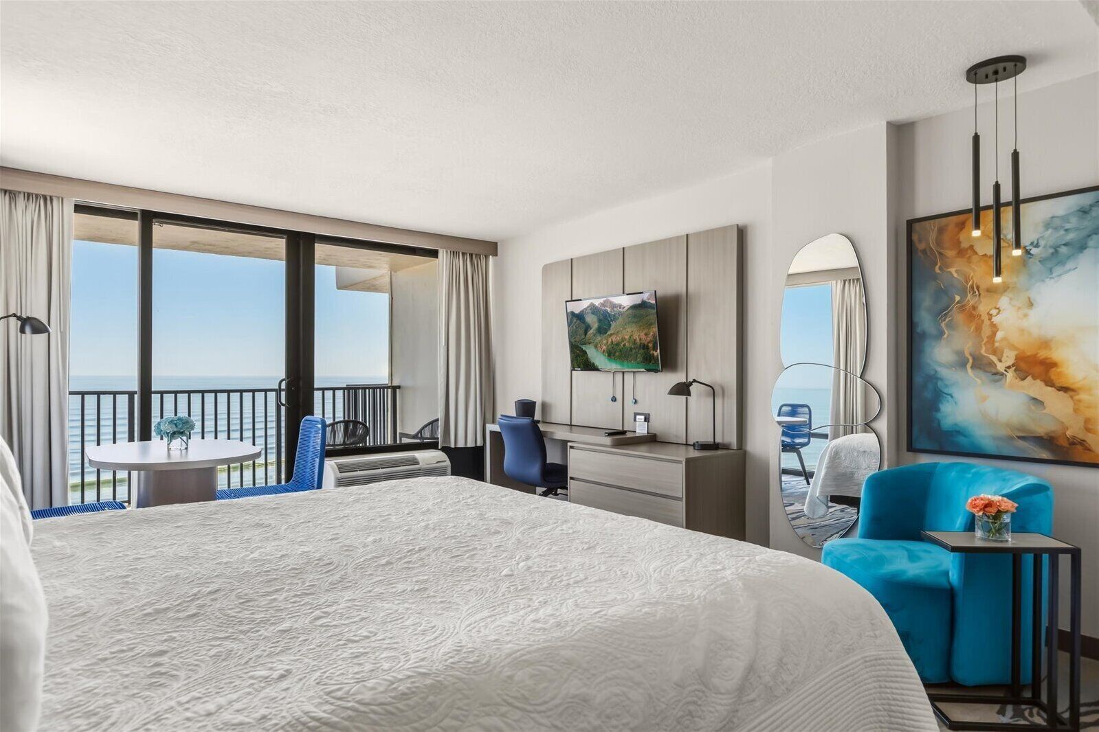 A hotel room with a large bed, a desk with a TV, a balcony with a table and chairs, and a painting on the wall.