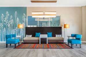 Une chambre d’hôtel moderne avec un grand lit, deux fauteuils bleus, une petite table blanche et un tapis coloré.
