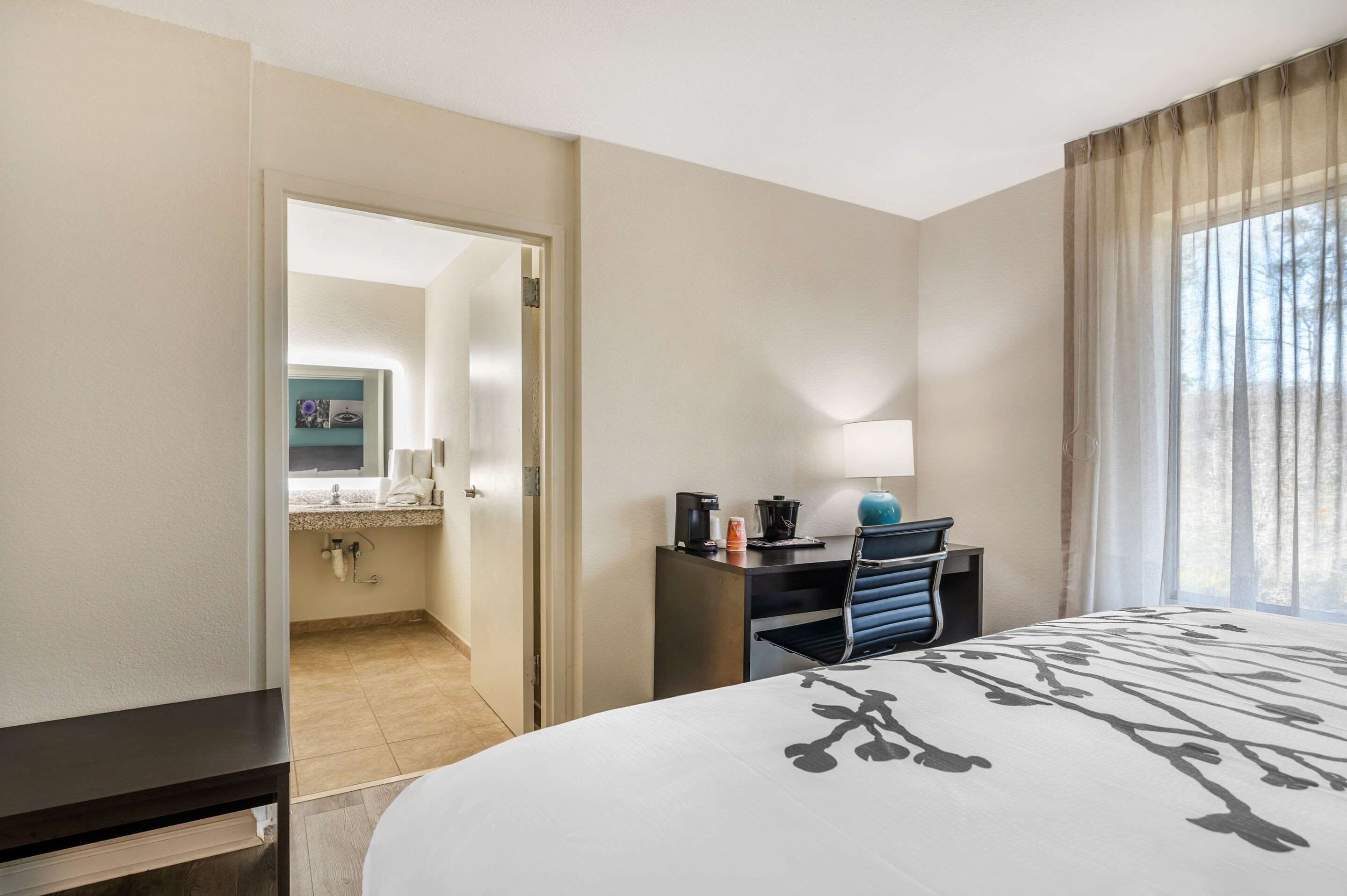 A hotel room with a bed, a desk with a lamp, a chair, and a view of the outdoors through a window.