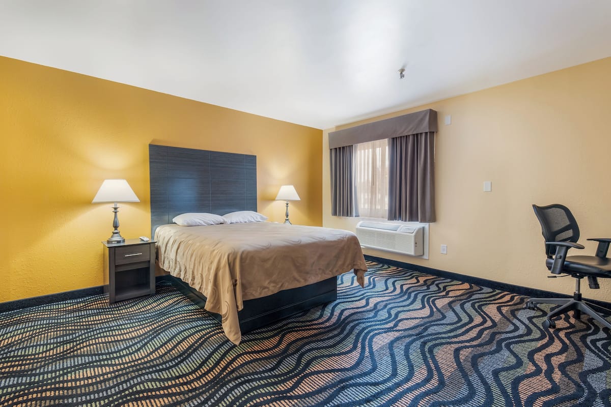 A hotel room with a bed, two bedside tables with lamps, a chair, a window with curtains, and a patterned carpet.