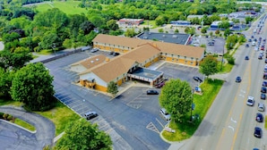 An aerial view of a commercial building with a parking lot, surrounded by greenery and a busy road.