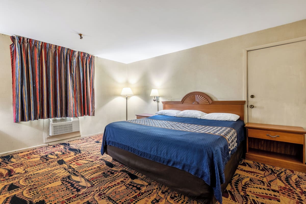 A hotel room with a bed, bedside lamps, a wooden nightstand, a patterned carpet, and a window with curtains.
