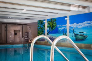An indoor swimming pool with clear poolside steps, a mural of a tropical beach scene, and a sauna area. - Hotel Park Soltau (Soltau)