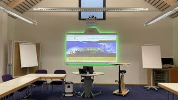 A conference room with a large projection screen displaying a website, a whiteboard, a table with chairs, and a ceiling with wooden beams.