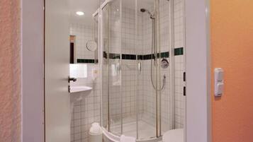 A glass-enclosed shower stall with a white sink and toilet, tiled walls, and a visible trash can.