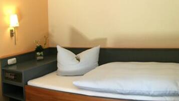 A neatly made bed with white bedding and pillows, a bedside table with a lamp, and a small plant on the nightstand.