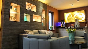 A modern living room with a grey sofa, a coffee table, and a decorative vase with flowers. There are built-in shelves with various items and a large window providing natural light. - Hotel Park Soltau (Soltau)
