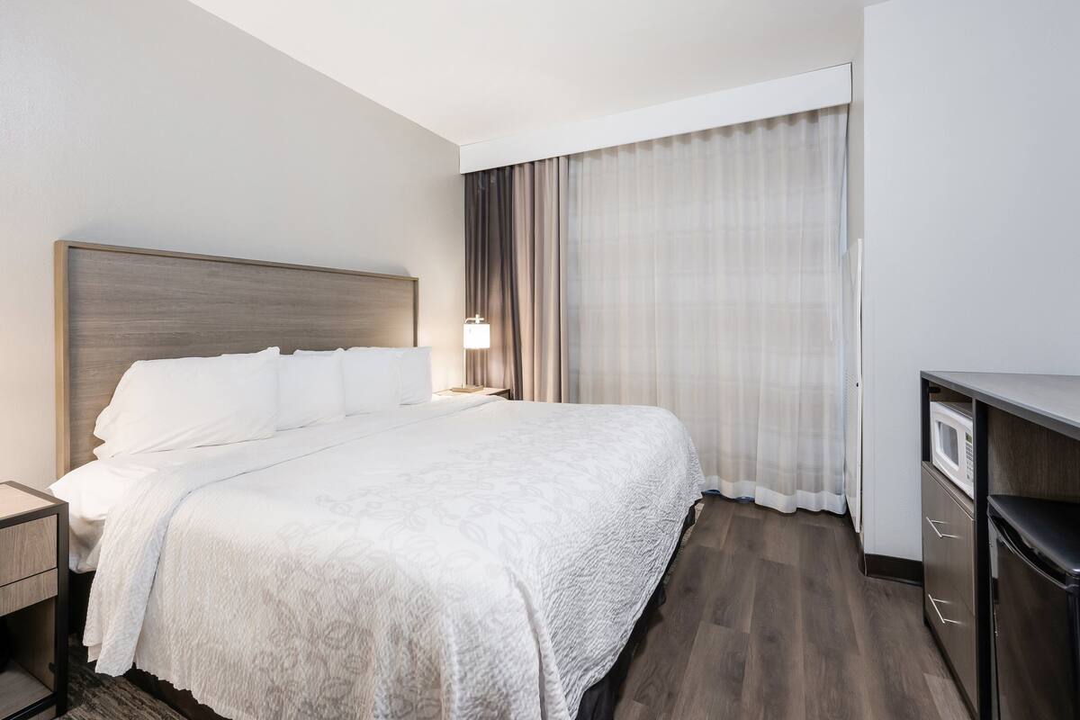 Habitación de hotel con una cama grande, una mesita de noche, una lámpara, un microondas y una puerta corredera con cortinas.