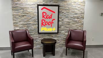 A Red Roof Plus sign on a stone wall, two leather chairs, and a small round table in a lobby area.