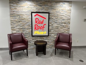 A Red Roof Plus sign on a stone wall, two leather chairs, and a small round table in a lobby area.