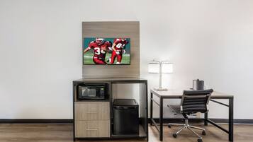 A hotel room with a flat-screen TV mounted on the wall, a microwave, a mini fridge, a desk with a chair, and a lamp.