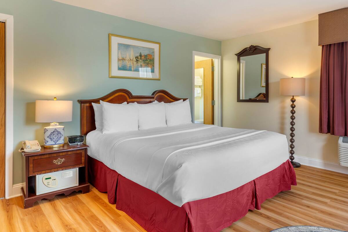 A neatly made bed with a white comforter and red skirt, a wooden nightstand with a lamp, a framed painting, and a mirror on the wall.