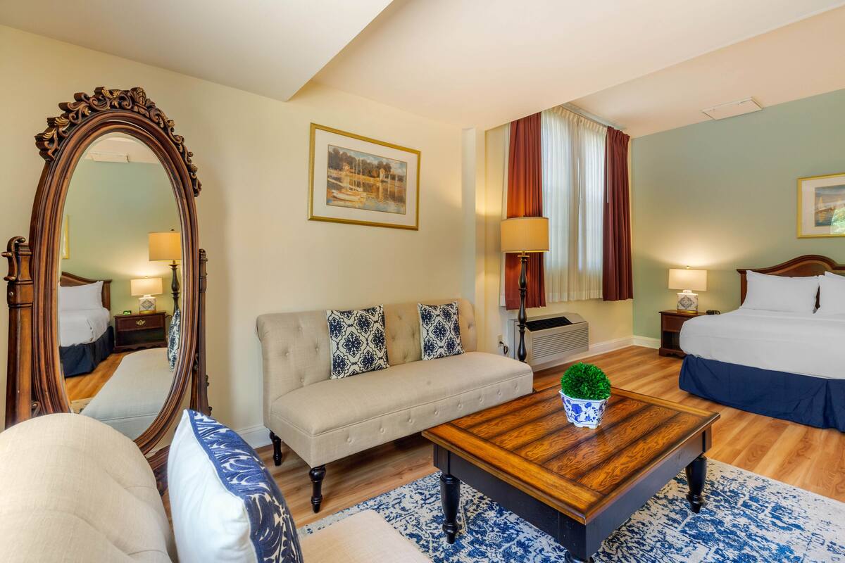 A hotel room with a large mirror, a sofa, a bed, a wooden coffee table, and a small potted plant.