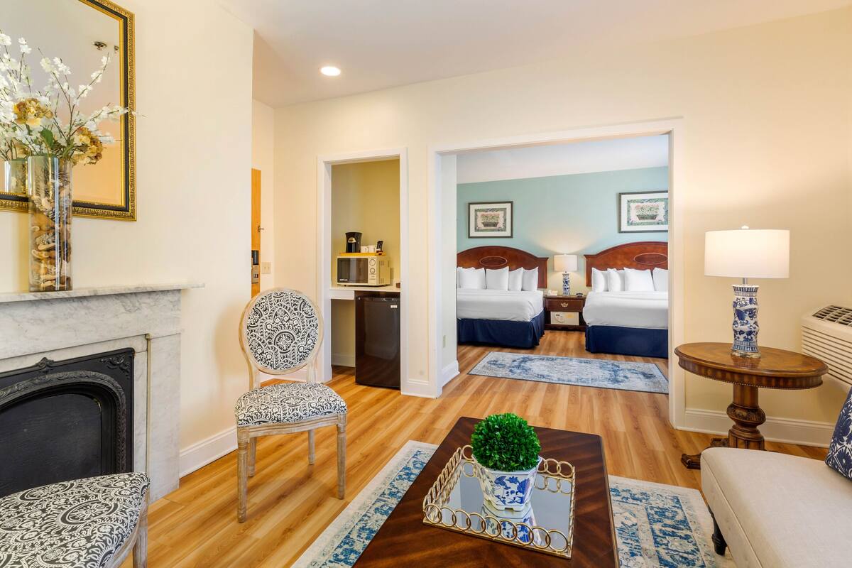 A hotel room with a fireplace, a dining table with a vase, a bed with a nightstand, and a bathroom visible through an open door.