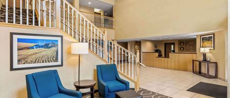 Un hall de hotel con una escalera, sillones azules, un mostrador de recepción y un cuadro enmarcado en la pared.