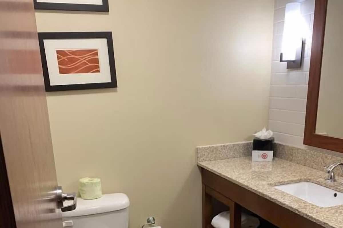 A bathroom with a toilet, sink, and mirror. There are framed pictures on the wall and a towel dispenser.