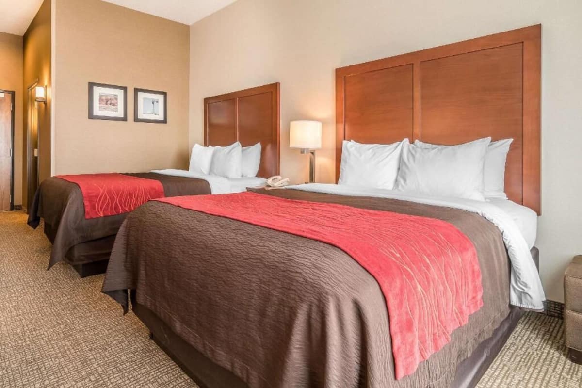 A hotel room with two beds, a wooden headboard, a nightstand with a lamp, and a carpeted floor.