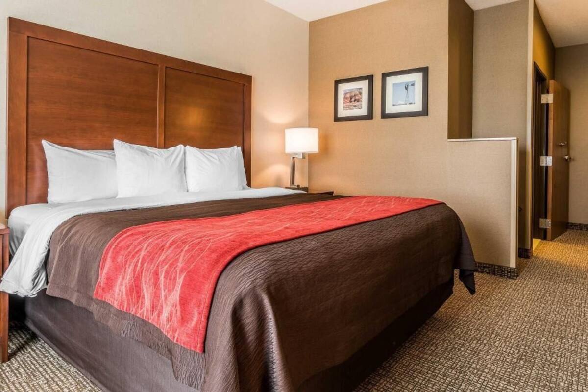A hotel room with a large bed, a nightstand, a lamp, a refrigerator, and a door leading to another room.
