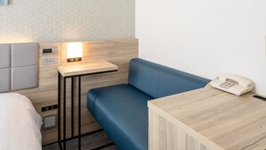 Une chambre d’hôtel compacte avec un canapé bleu, une table de chevet en bois avec un téléphone et une petite table de chevet avec une horloge numérique.