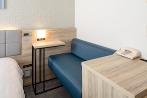 Ein kompaktes Hotelzimmer mit einem blauen Sofa, einem Nachttisch aus Holz mit einem Telefon und einem kleinen Nachttisch mit einer digitalen Uhr.