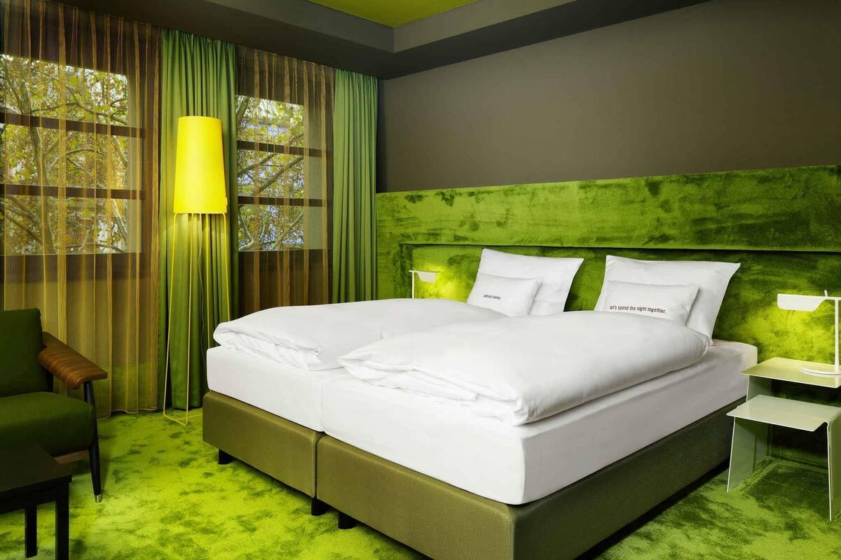 Une chambre d’hôtel avec un grand lit, une moquette verte, un fauteuil vert, une table de chevet et une fenêtre avec des rideaux.