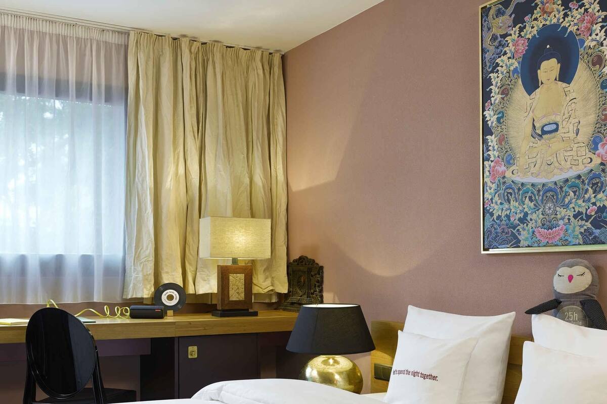 Une chambre d’hôtel comprenant un lit, deux tables de chevet avec des lampes, un bureau avec une chaise, une fenêtre avec des rideaux et un mur orné d’un tableau encadré.