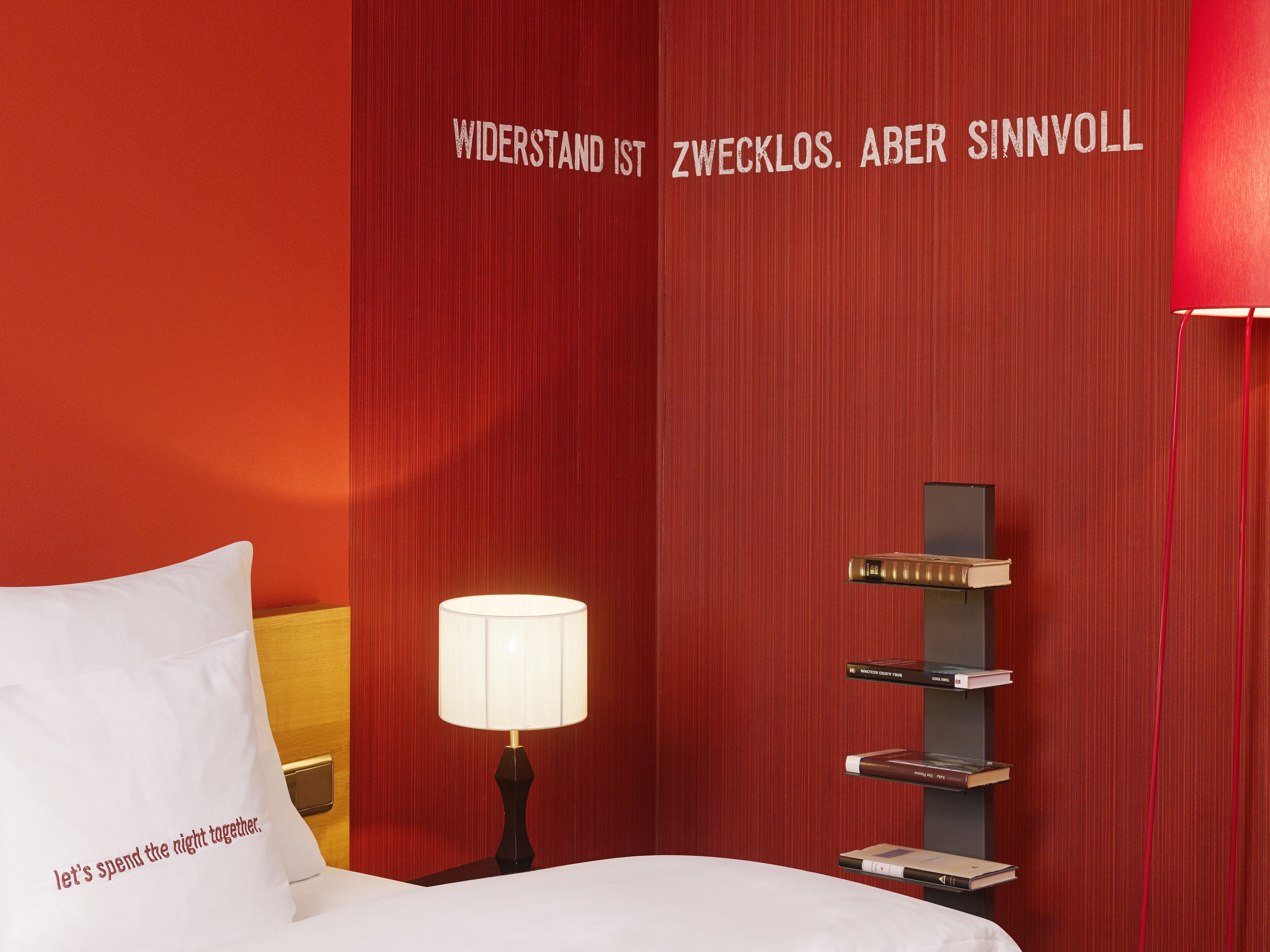 A hotel room with a red wall, a bedside table with a lamp, a bookshelf, and a pillow with text.