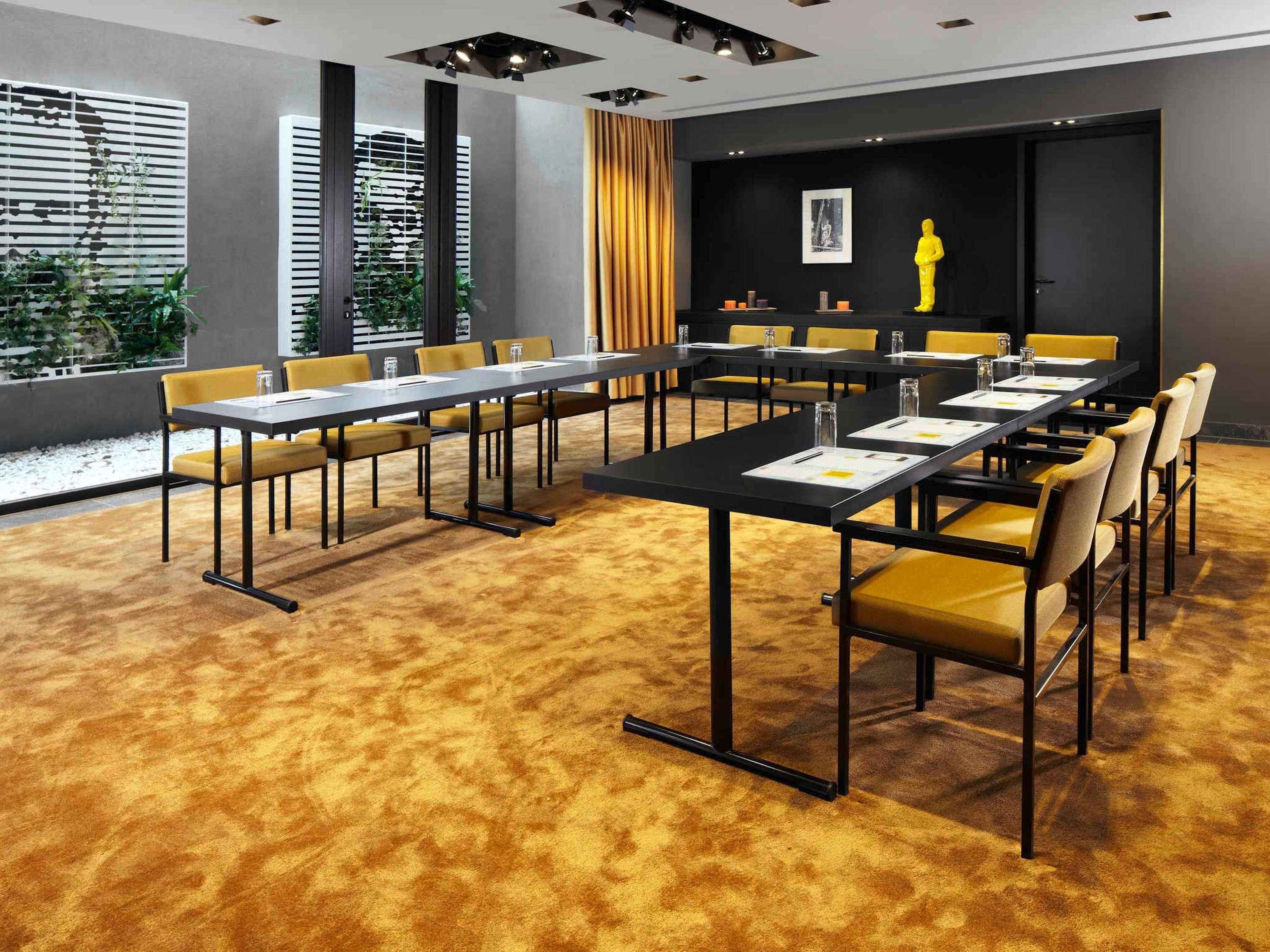 Een vergaderruimte met meerdere tafels en stoelen, een groot raam met zonwering en een geel gordijn.