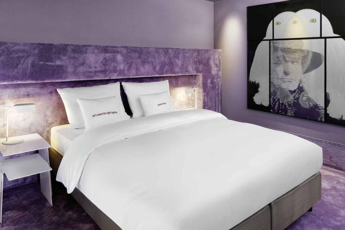 Une chambre moderne avec un grand lit, du linge de lit blanc et un mur d’accent violet.