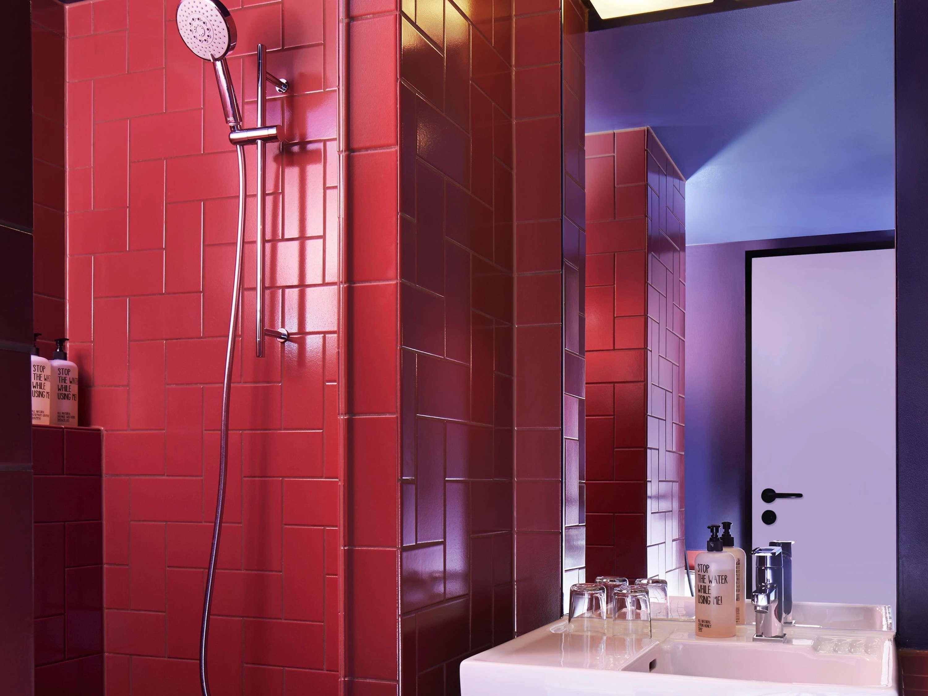 Un baño con paredes de azulejos rojos, ducha, lavamanos con espejo y artículos de aseo.