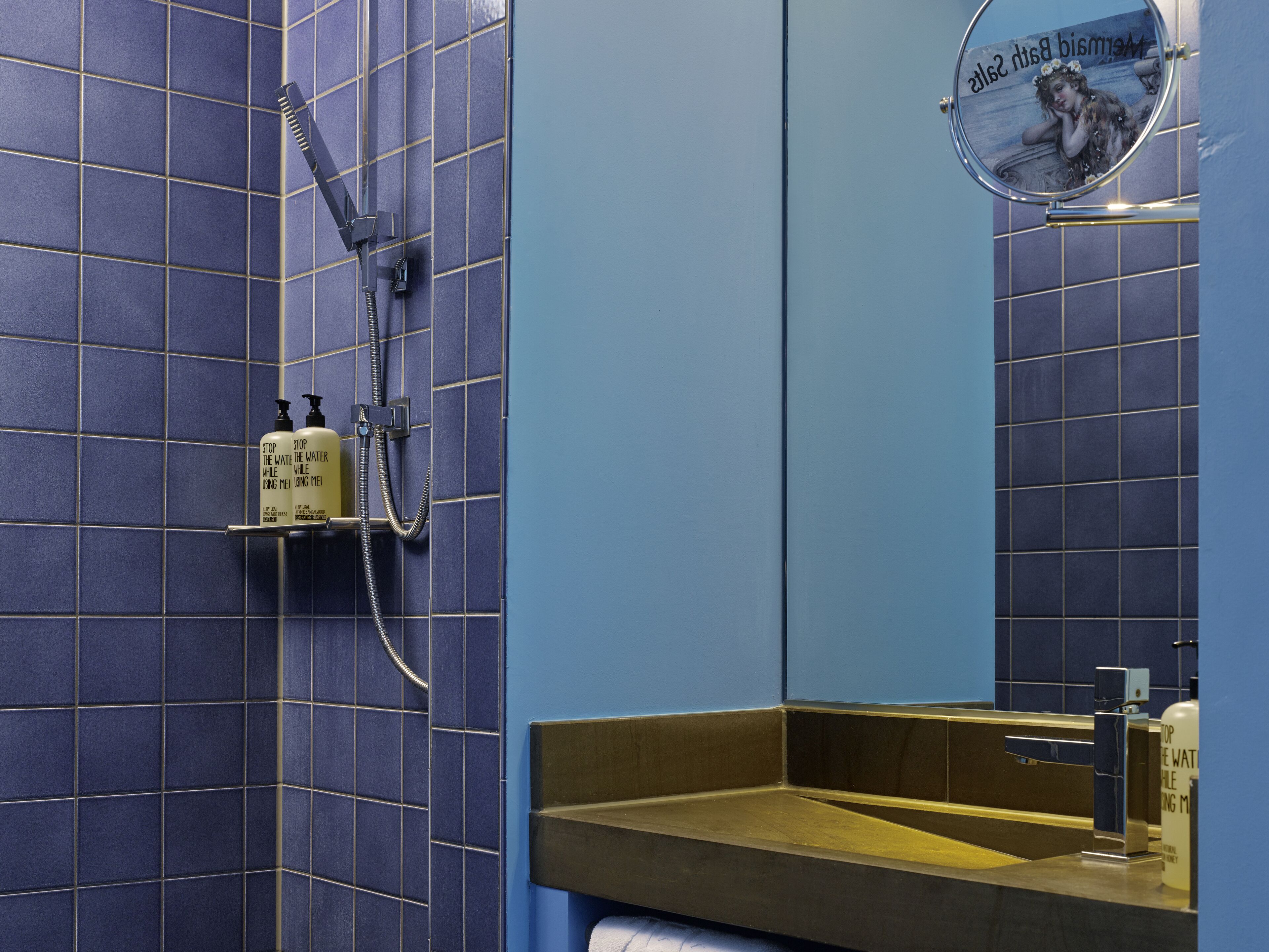 A bathroom with blue tiled walls, a shower, a sink with a mirror, and two bottles of shampoo.