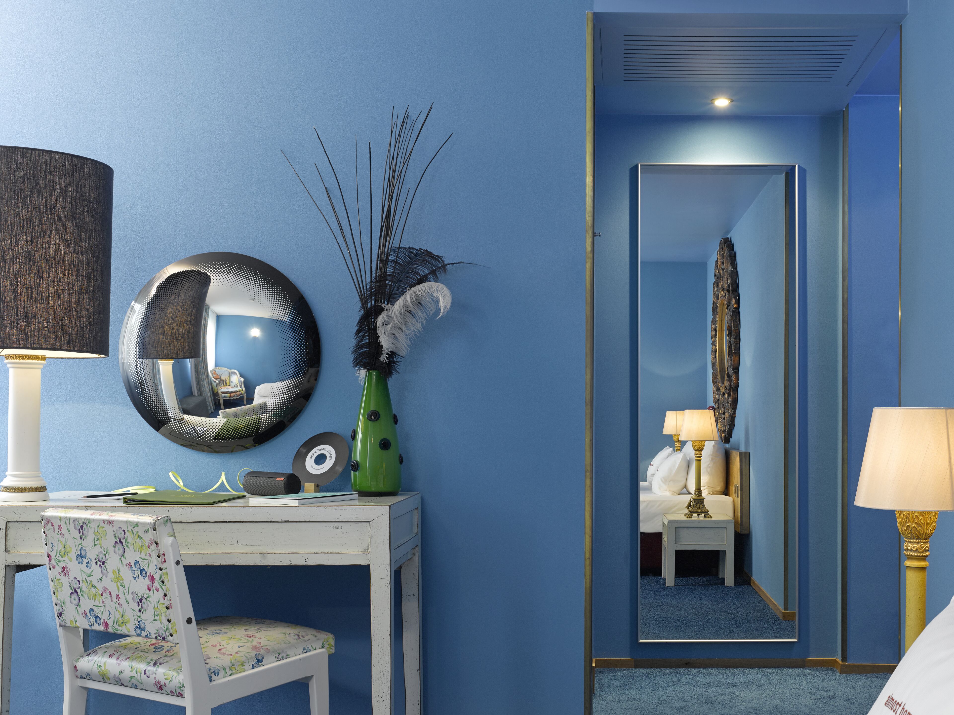Une pièce avec un mur bleu, un miroir rond, un bureau avec une chaise et un lit avec une table de chevet.