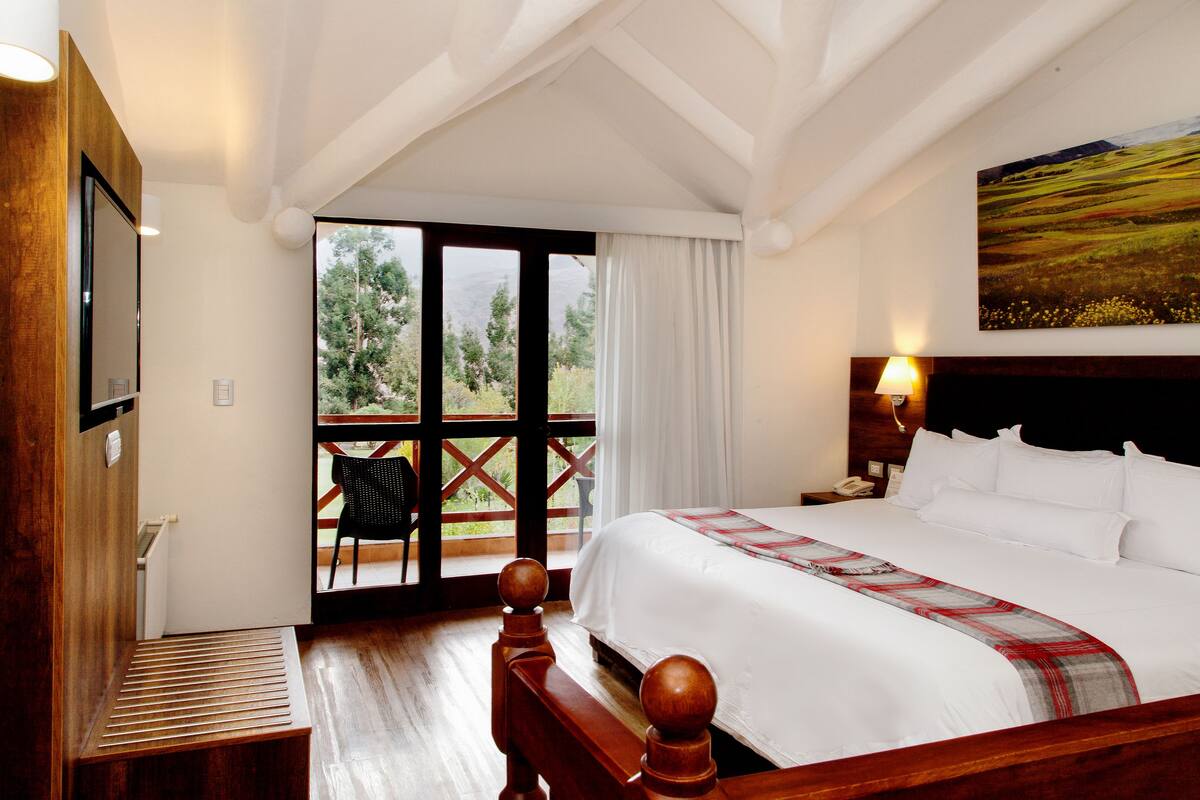 Une chambre avec un grand lit, une tête de lit en bois, une table de chevet, une lampe et une vue sur un espace extérieur aménagé avec une chaise, accessible par une porte vitrée.