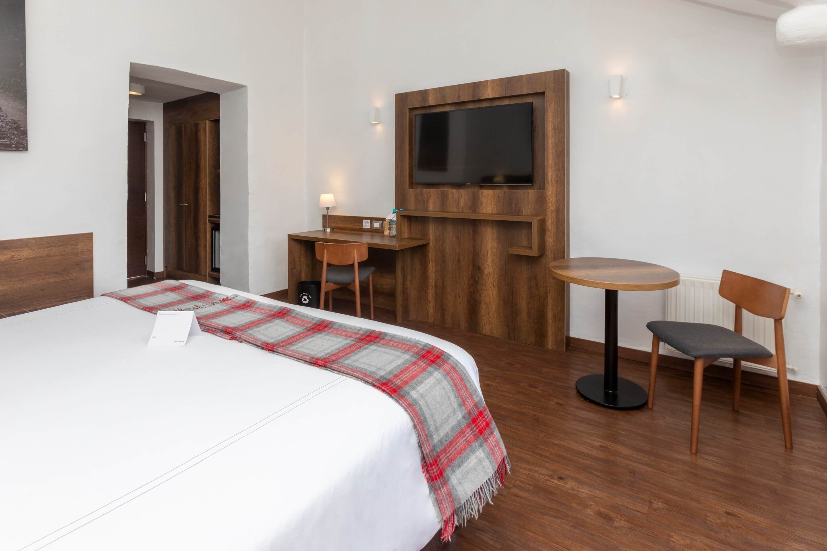 A hotel room with a large bed, a desk with a chair, a small round table with a chair, and a flat-screen TV mounted on the wall.