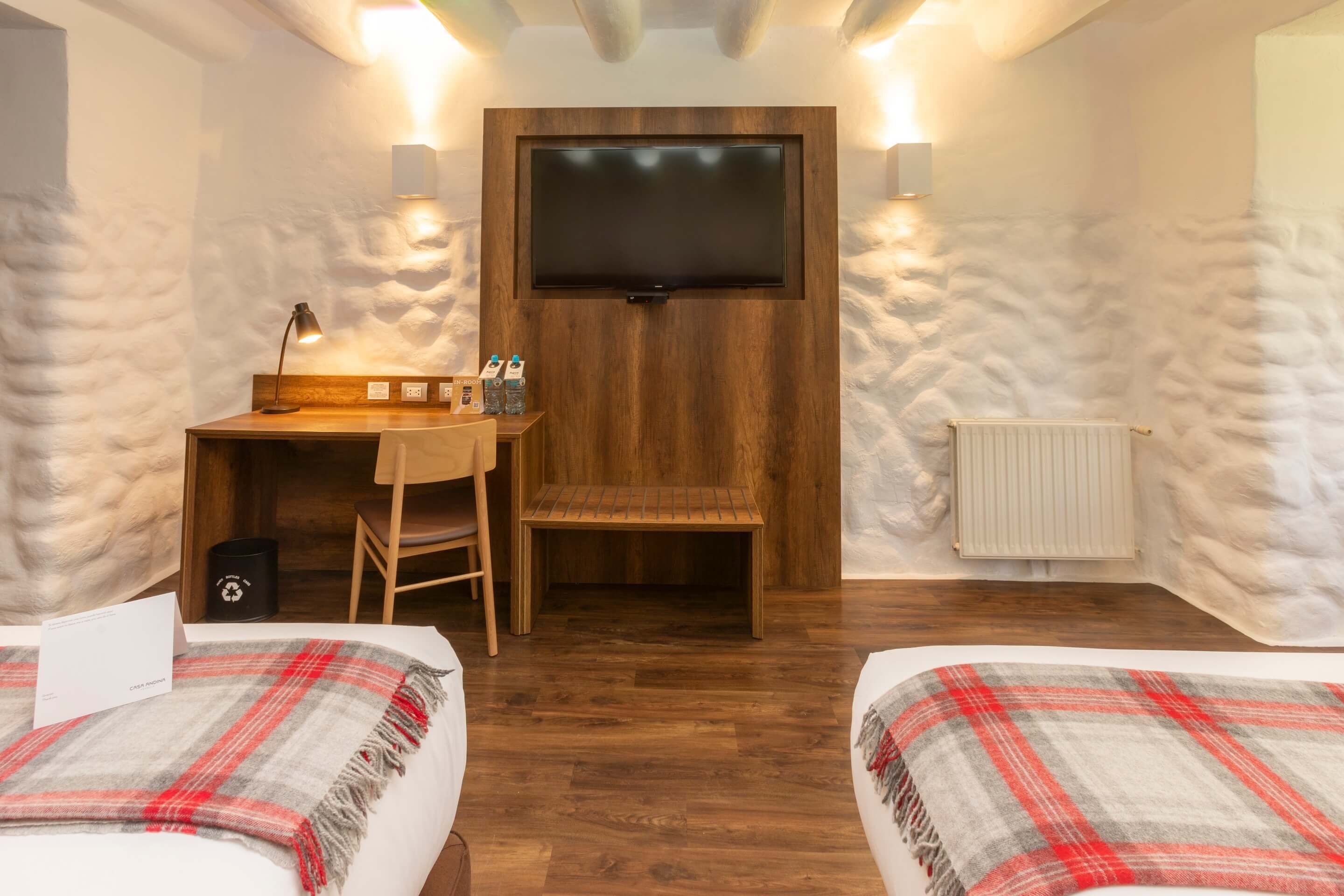 A cozy room with a wooden desk, a TV mounted on the wall, and two beds with plaid blankets.