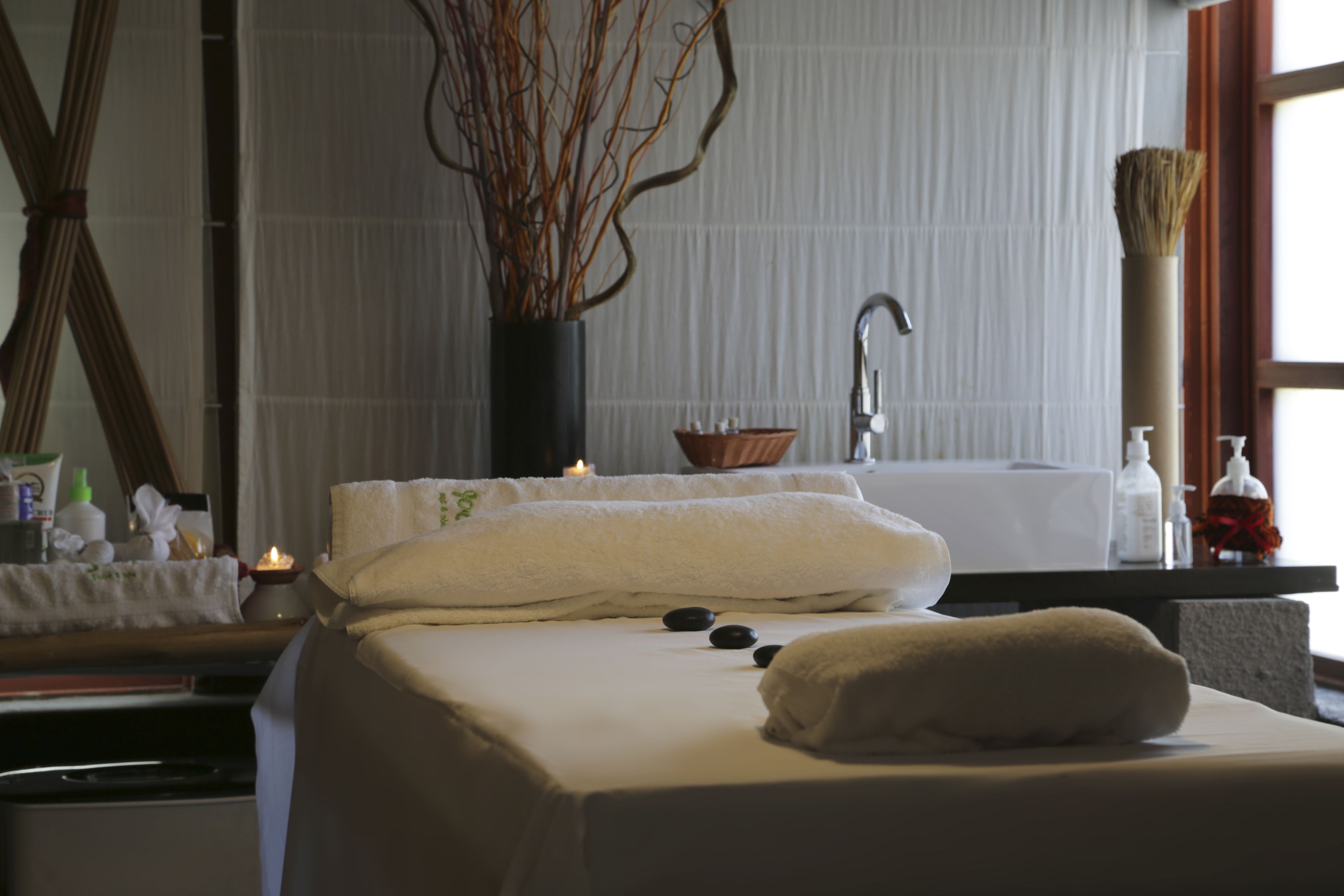 A spa room with a massage table, candles, a vase with branches, and a bathtub.