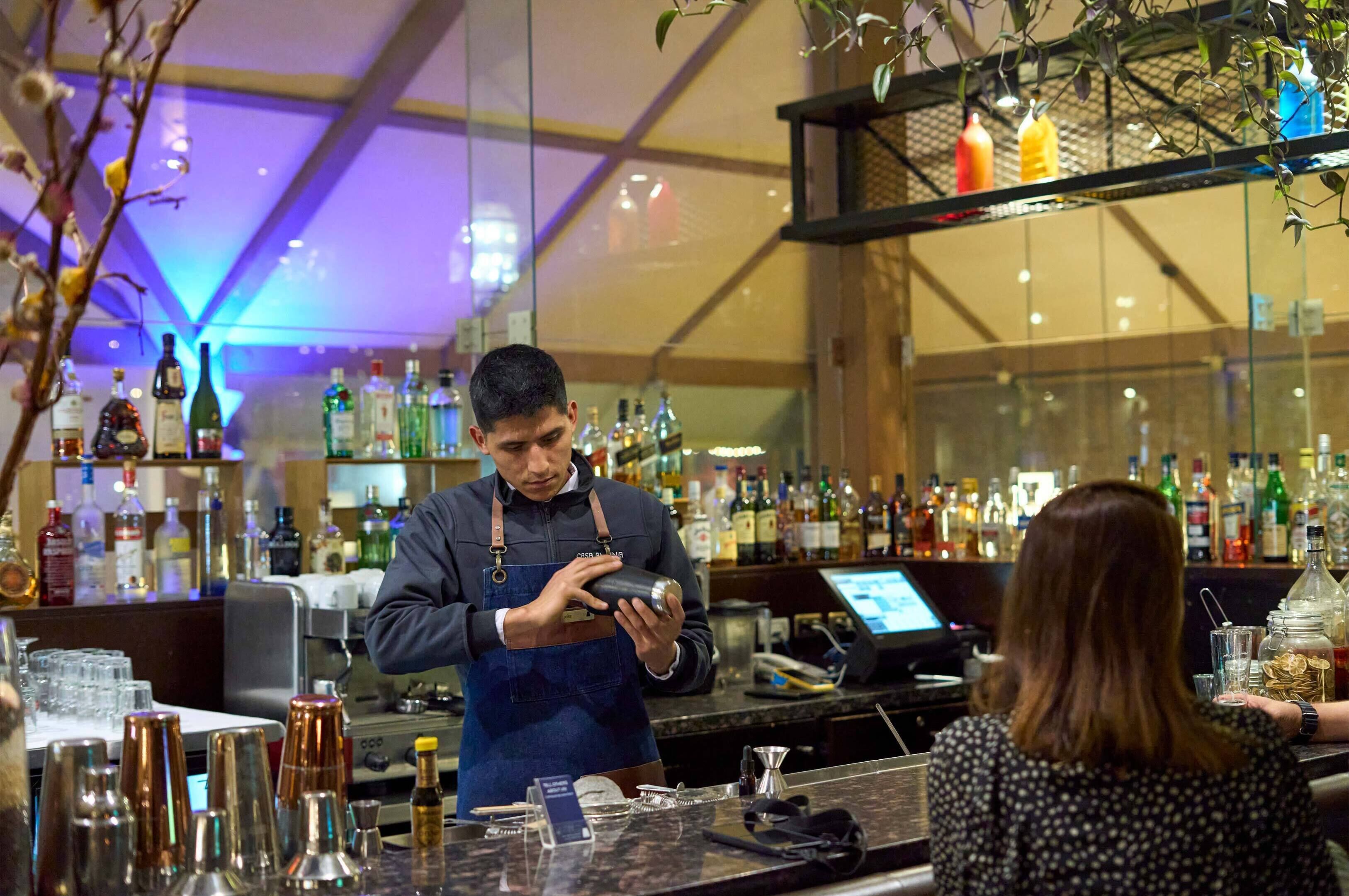 Een bar met een bartender die een drankje klaarmaakt, een klant zittend, en verschillende flessen en barbenodigdheden op de toonbank.
