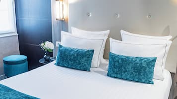 A neatly made bed with white linens and two teal pillows, a small bedside table with a vase of flowers, and a blue ottoman.