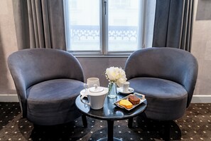 Ein Zimmer mit zwei grauen Sesseln, einem kleinen runden Tisch mit einem weißen Teekessel, Tassen, einer Vase mit weißen Blumen und einem Tablett mit zwei Gebäckstücken.