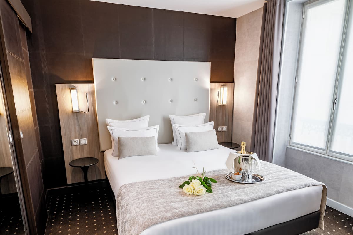 Une chambre d’hôtel avec un grand lit, deux tables de chevet, une petite table, une fenêtre avec des rideaux et un luminaire fixé au mur.