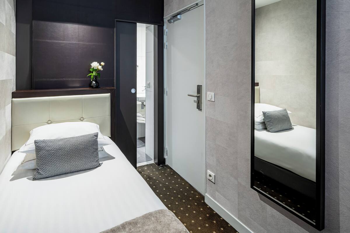 Une chambre d’hôtel moderne, équipée d’un lit, d’une table de chevet ornée d’un vase de fleurs, d’un grand miroir et d’une porte donnant sur une autre pièce.