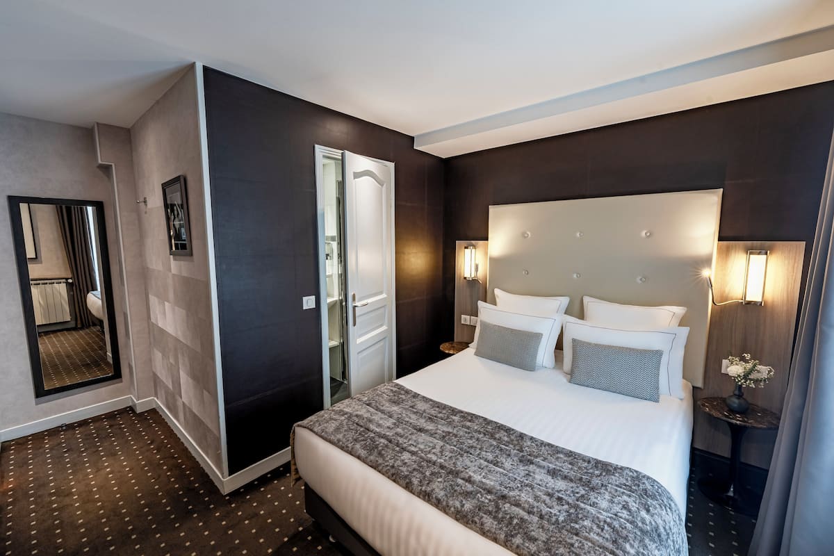 Une chambre d’hôtel avec un grand lit, une table de chevet avec une lampe, un miroir et une porte donnant sur une salle de bain.