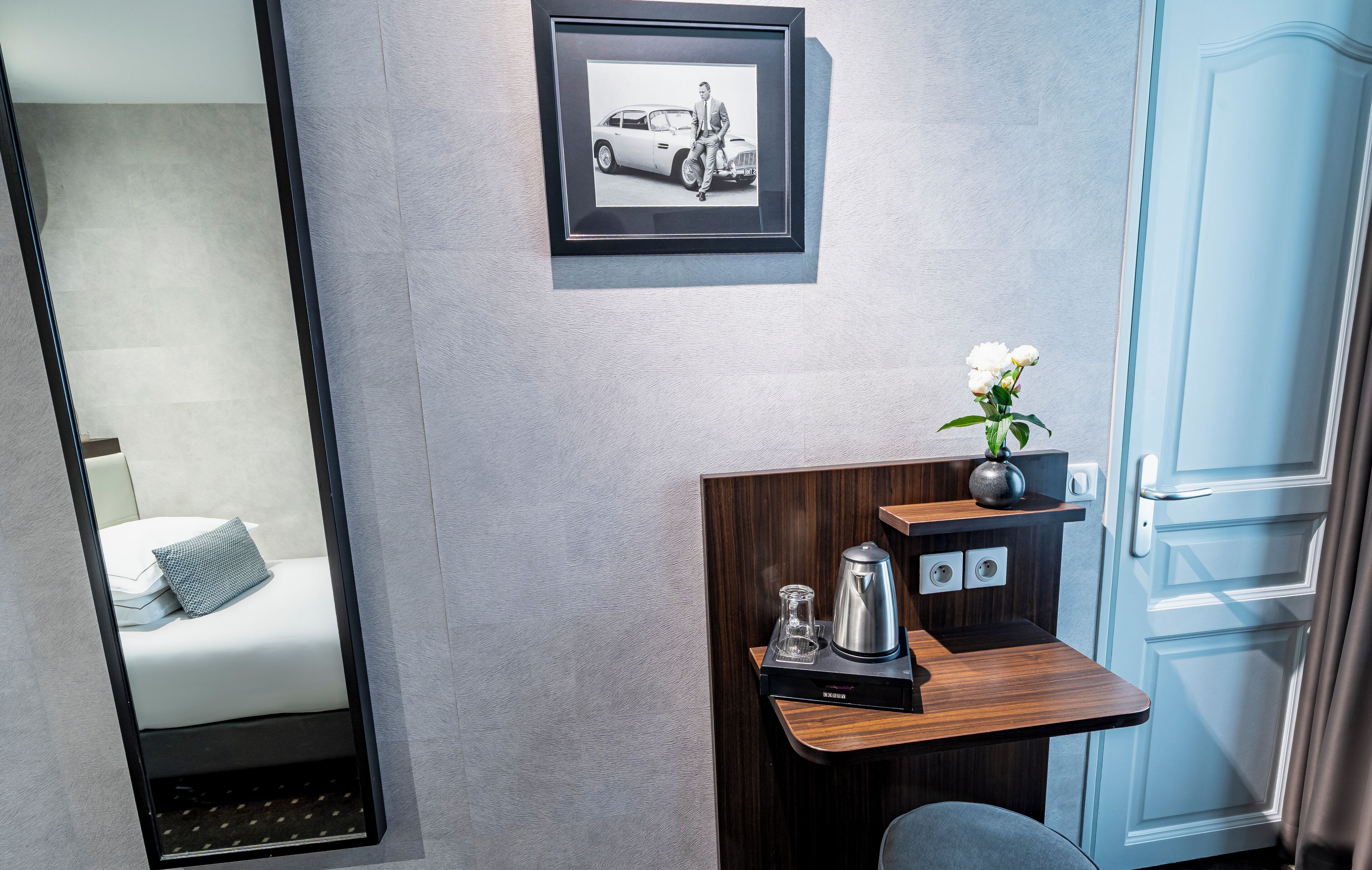A hotel room with a bed, a mirror, a framed picture of a car, a small table with a vase of flowers, and a coffee maker.