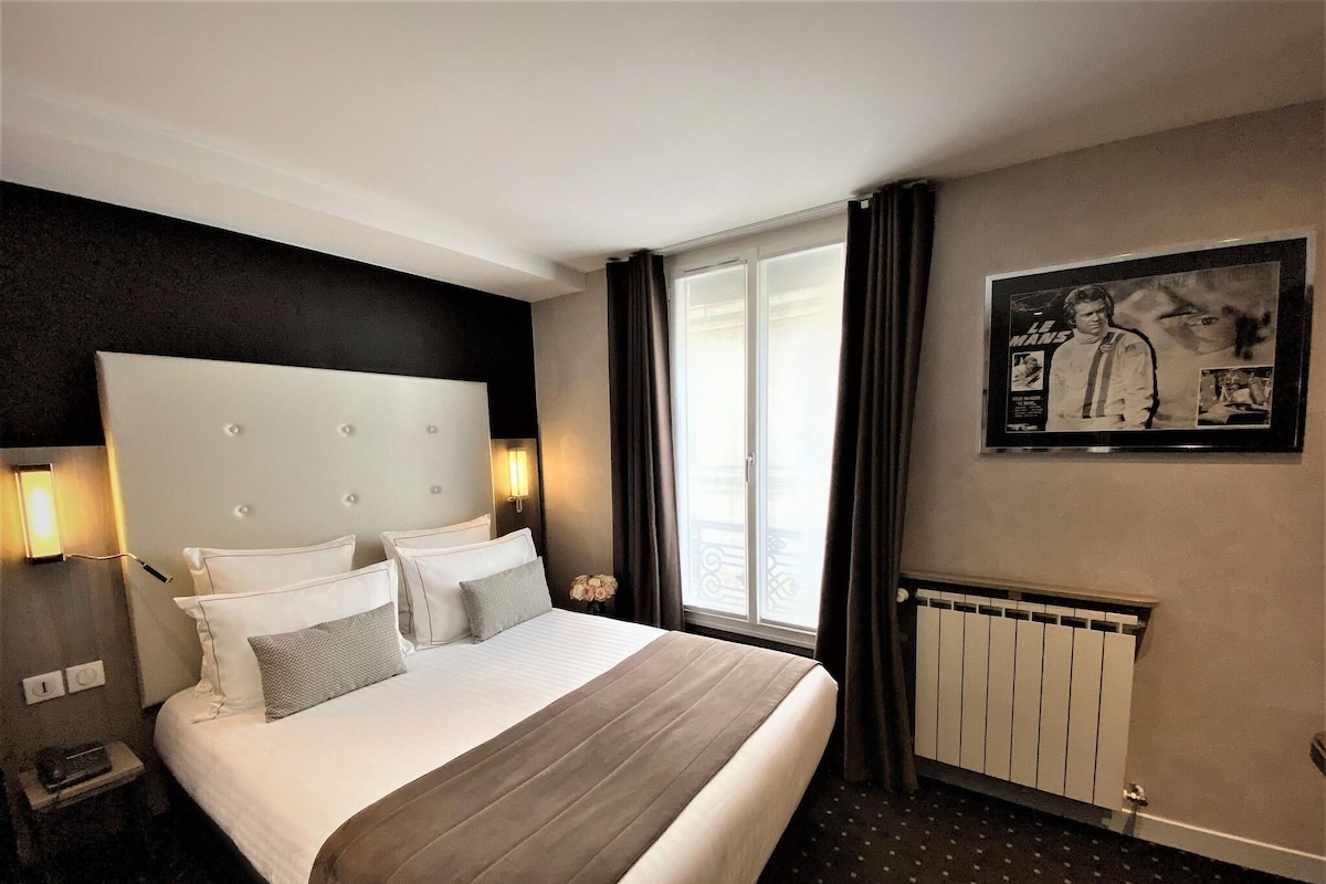 A hotel room with a large bed, a headboard, a nightstand, a lamp, a window with curtains, a radiator, and a framed picture on the wall.