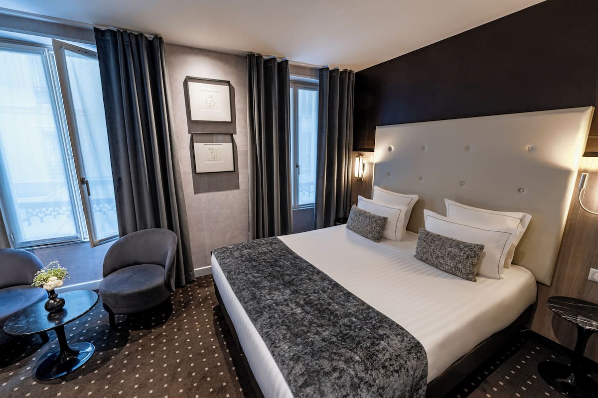 Une chambre d’hôtel avec un grand lit, deux fauteuils, une petite table avec un vase, et une vue sur la ville grâce à de grandes fenêtres.
