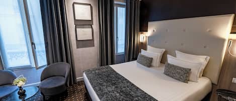 Habitación de hotel con una cama grande, dos sillones, una mesita con un florero y vistas a la ciudad a través de amplios ventanales.