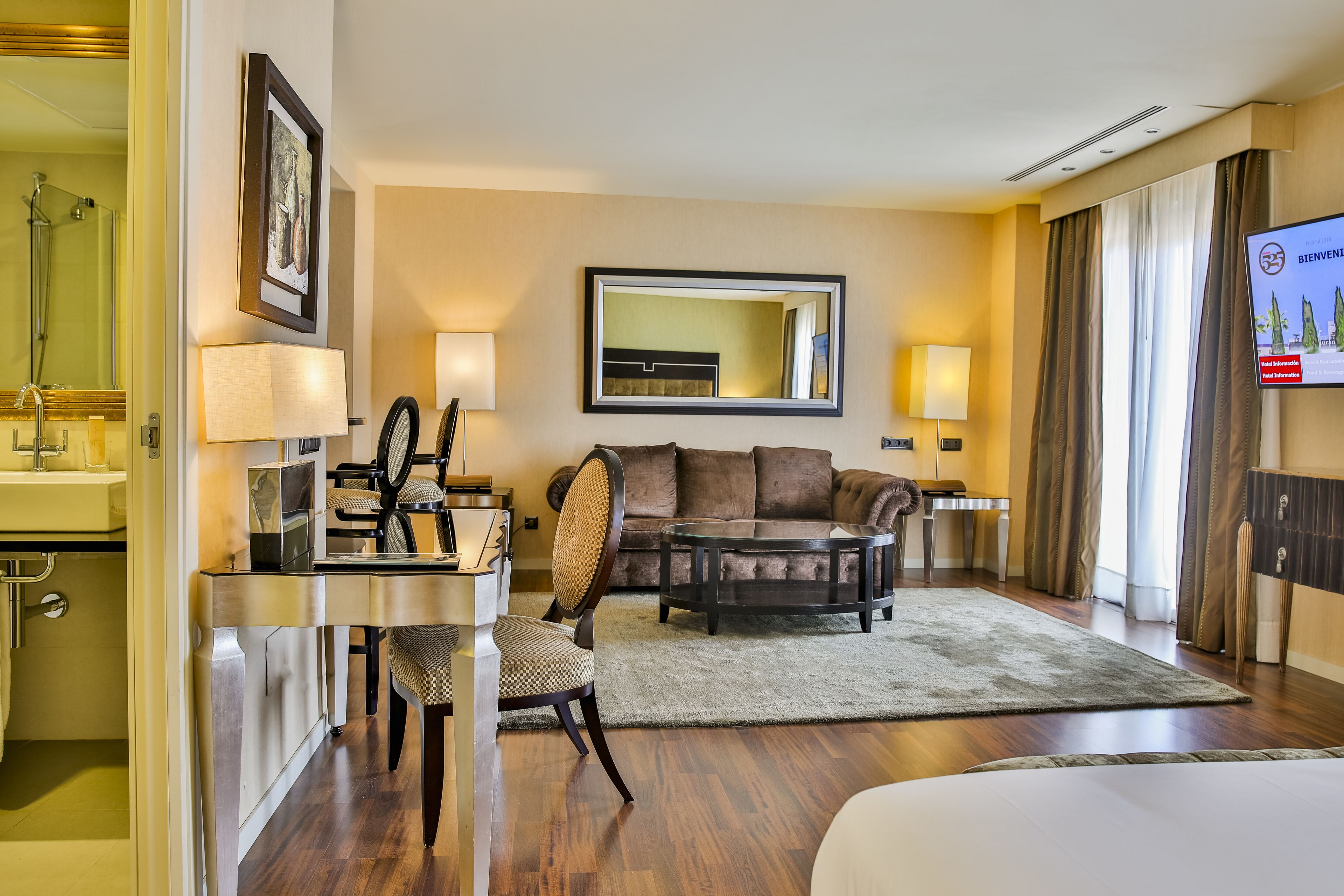 Una habitación de hotel con una cama, un escritorio con una silla, un sofá, una mesa de centro, un televisor y un baño visible a través de una puerta abierta.